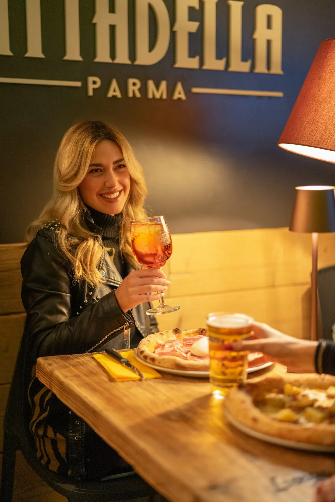Una ragazza sorride con uno spritz in mano davanti all’insegna Cittadella Parma